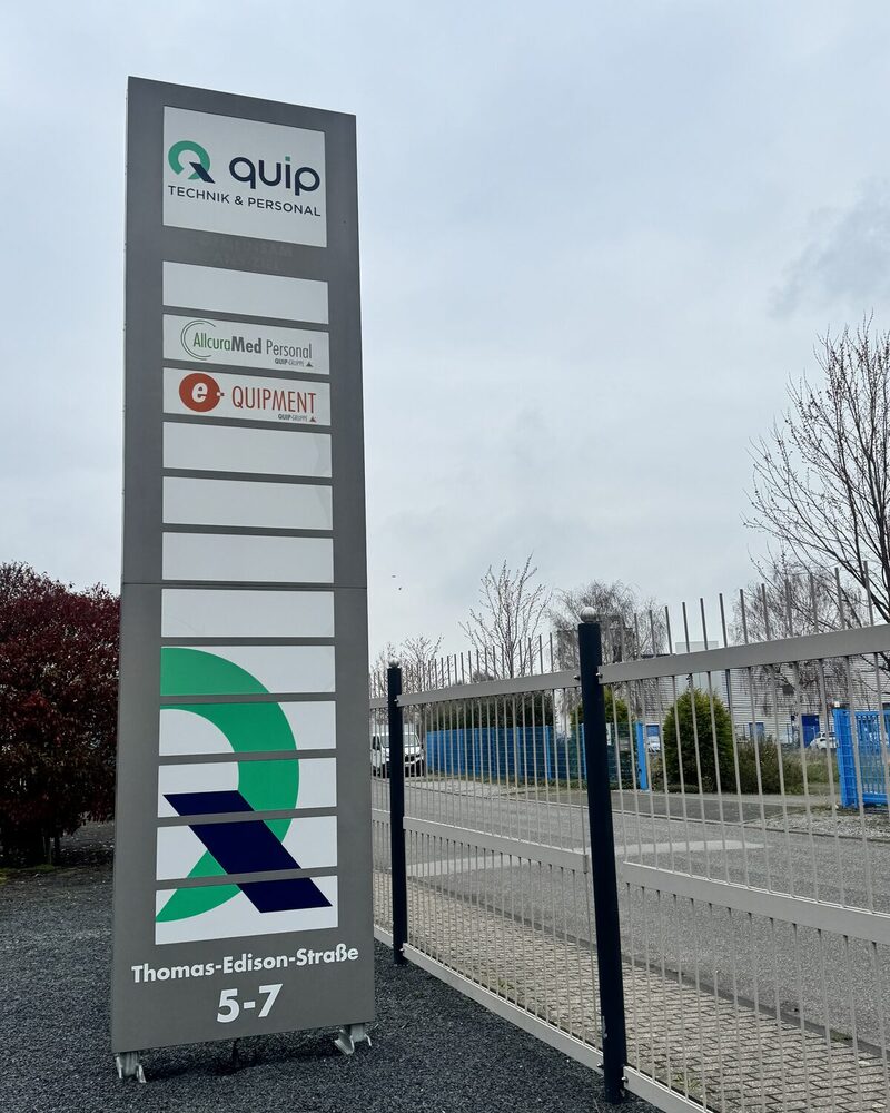 Firmenschild an der Zufahrt Thomas-Edison-Straße 5–7 mit den Schildern von quip, AllcuraMed Personal und e-QUIPMENT untereinander.