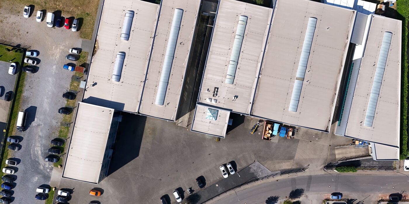 Senkrechte Luftaufnahme des Firmengeländes an der Thomas-Edison-Straße in Baesweiler: fünf Hallen mit Sheddächern, Parkplätze und die Zufahrt.